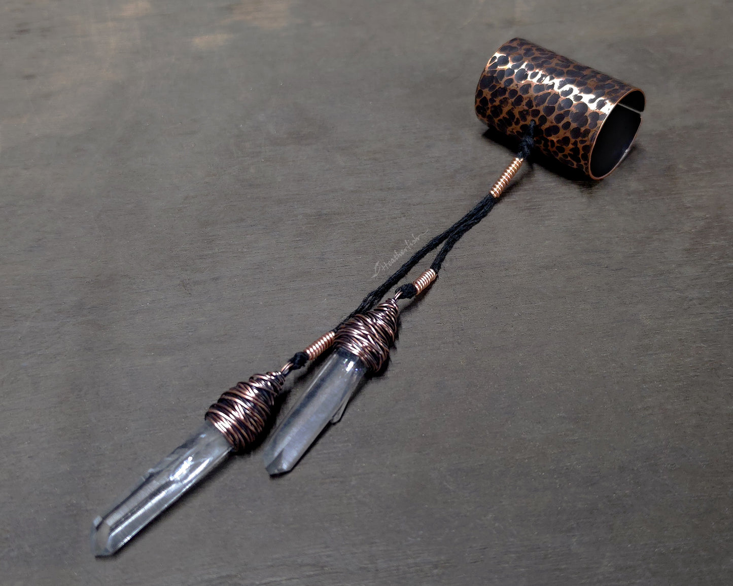 A handmade jewelry piece featuring two clear quartz crystal points wrapped in copper wire, connected by a black cord to a hammered copper cuff displayed on a dark gray textured surface.