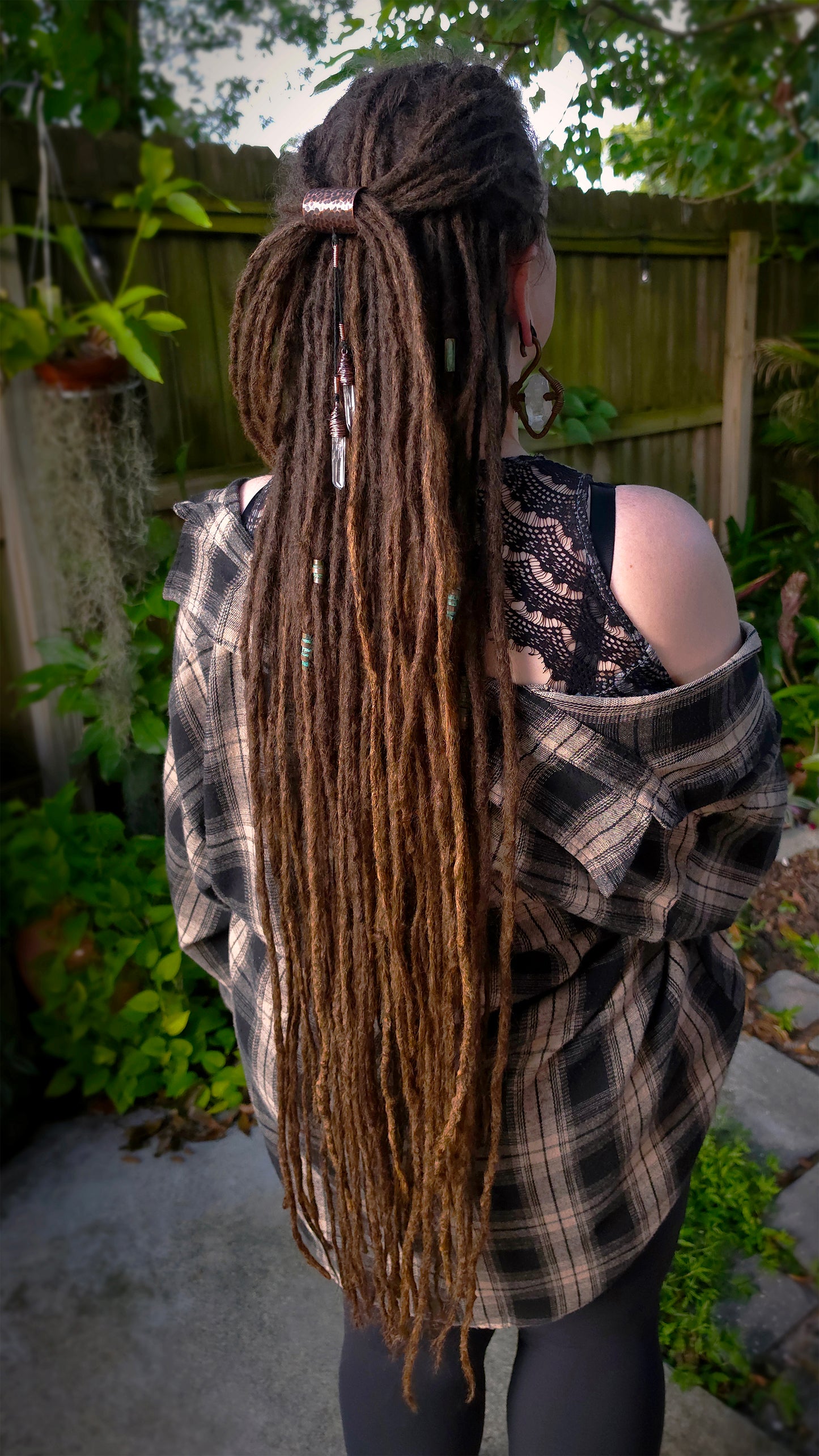 A person shown from the back outdoors, wearing a long brown dreadlock hairstyle with a half-up top knot and a small hair accessory. The person is wearing a copper ear weight, and an off-the-shoulder plaid flannel shirt over a black lace top, set against a backyard fence and garden background.