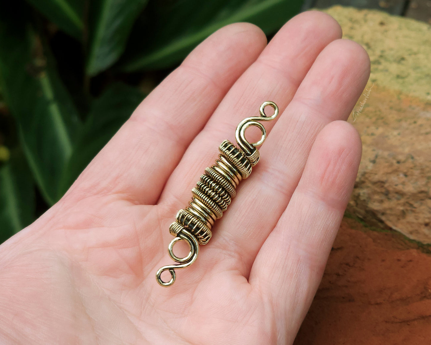 Woven brass dreadlock bead held in hand to show scale.