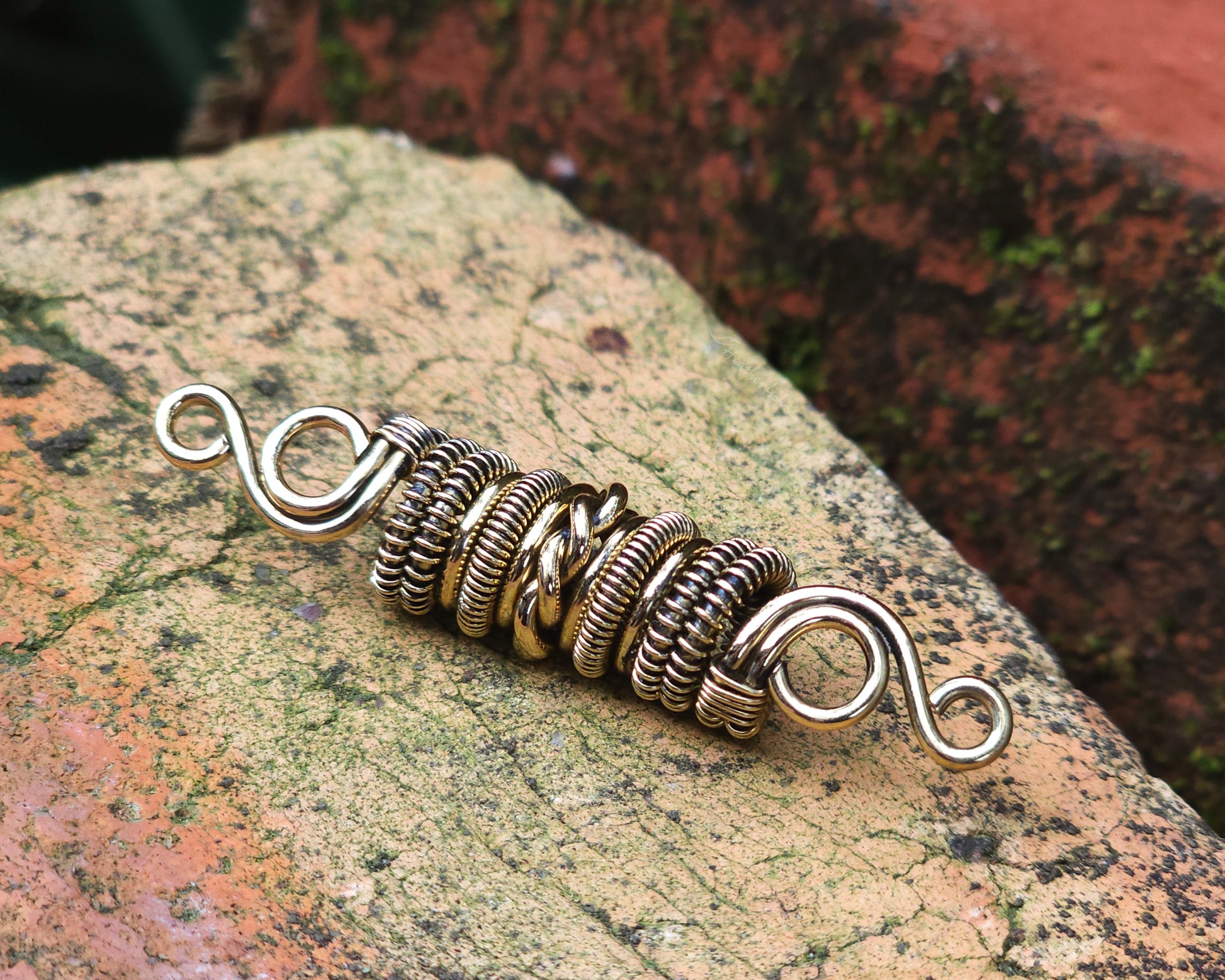 Woven brass dreadlock bead on a brick background.