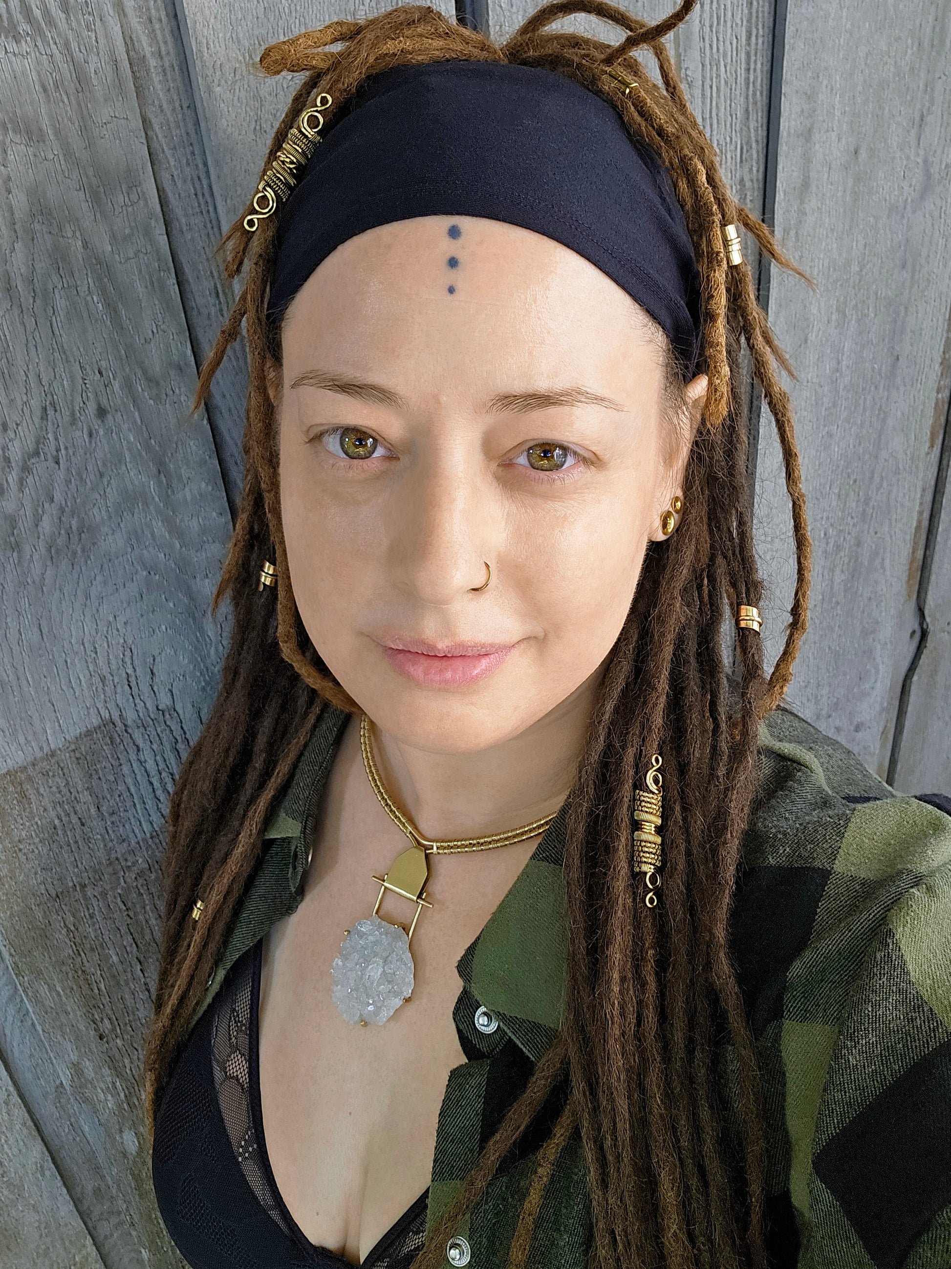 Person with long dreadlocks wearing a black headband, gold crystal necklace, and green shirt against a wooden background.