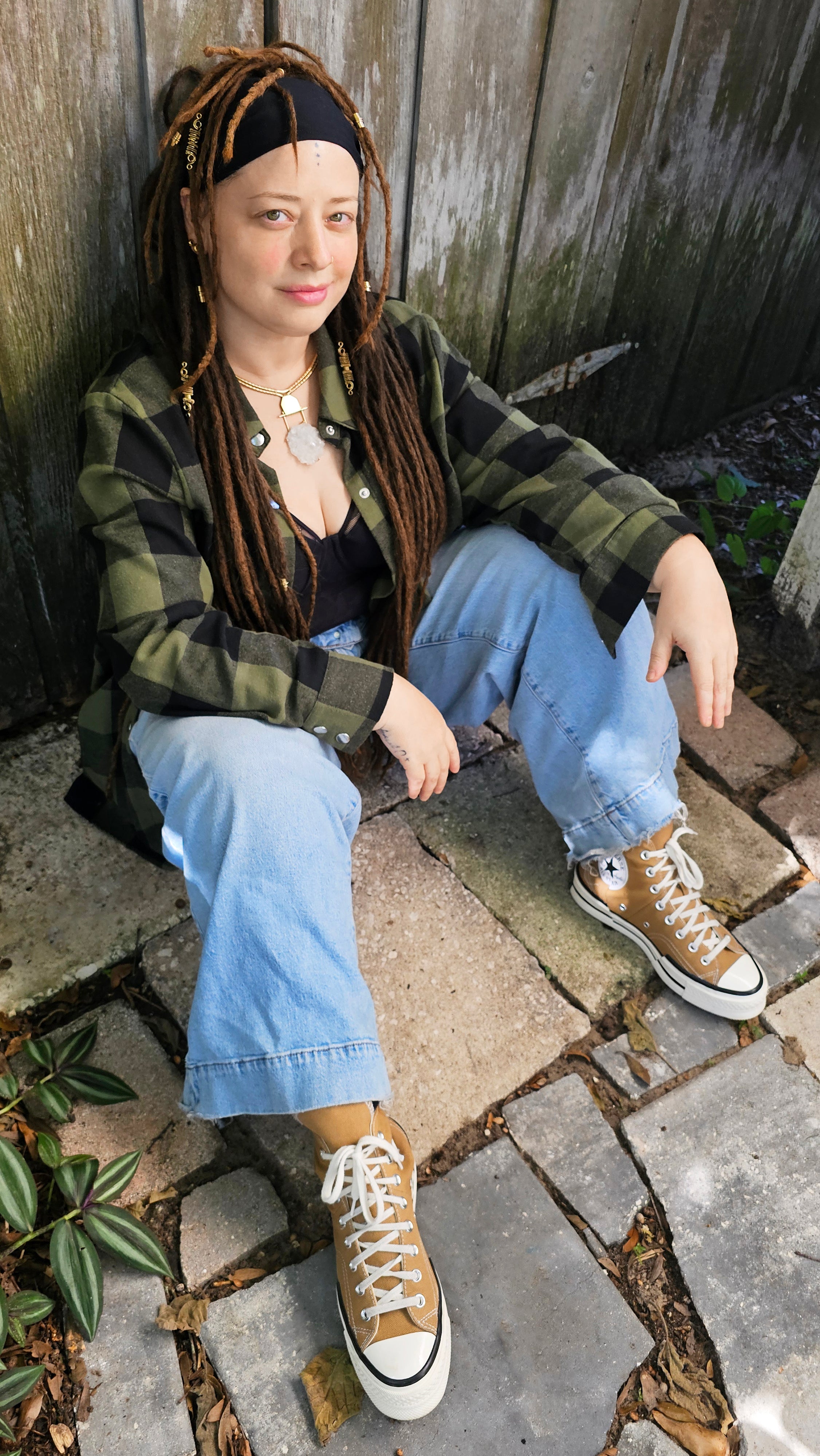 Person sitting on a stone path wearing a plaid shirt, gold crystal necklace, blue jeans, and brown converse sneakers.