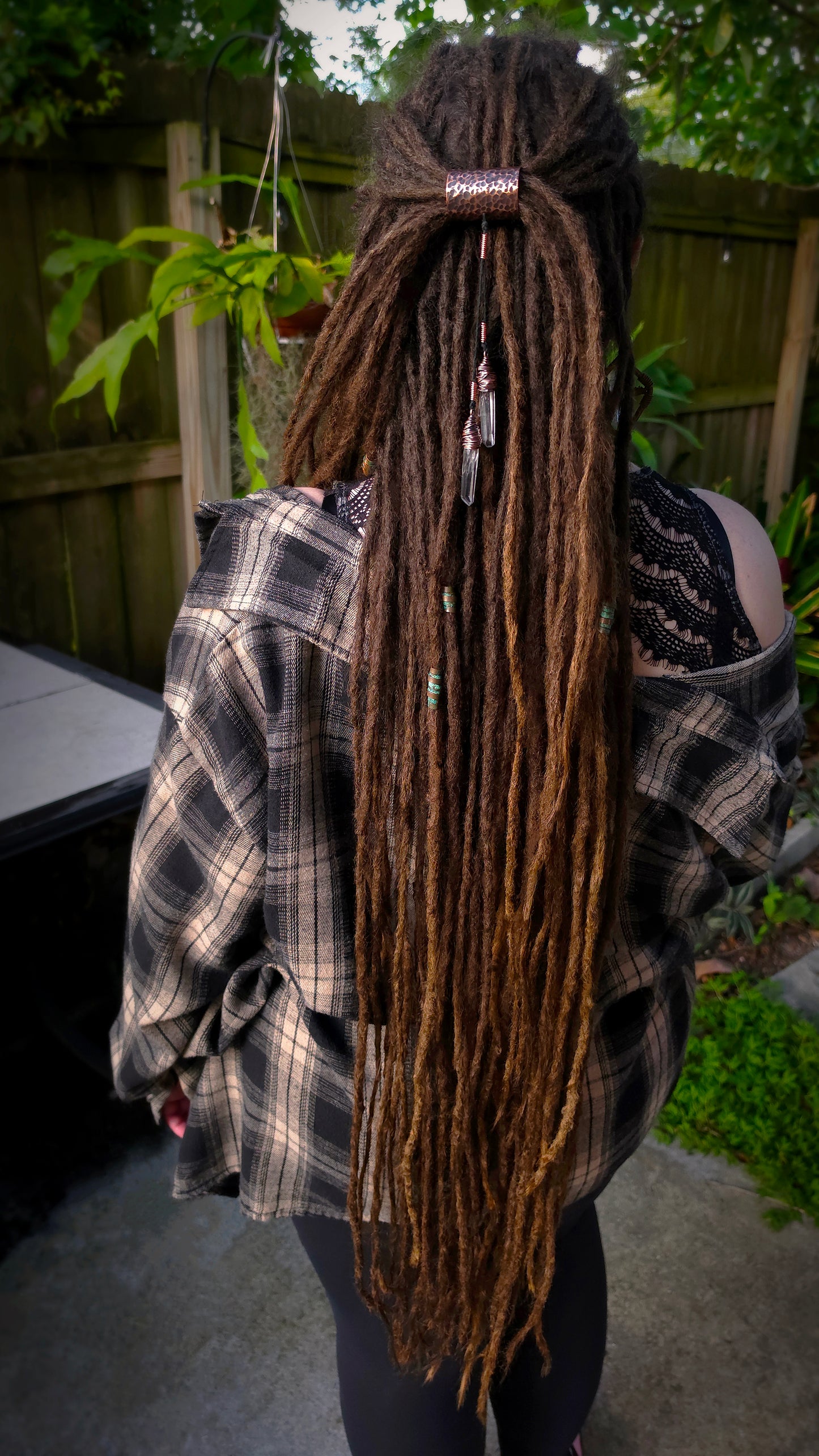 A person viewed from behind wearing a black and gray plaid flannel shirt over a black lace top, showcasing long, dark brown dreadlocks with small beads and a copper hair cuff. The setting is an outdoor backyard with a wooden fence and green plants.