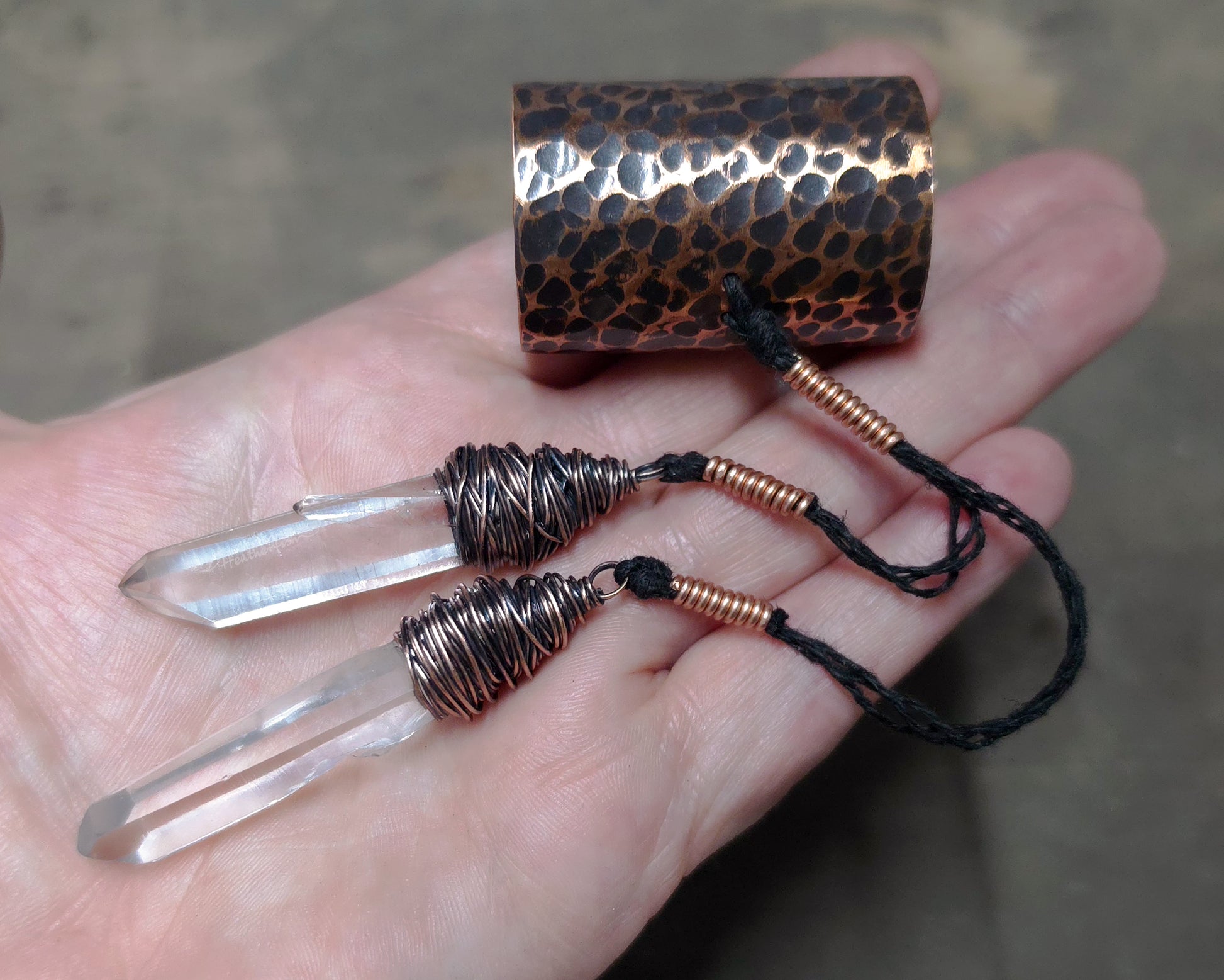 A hand holding a hammered copper hair cuff with two clear crystal points wrapped in copper wire, and black cords against a blurred gray background.