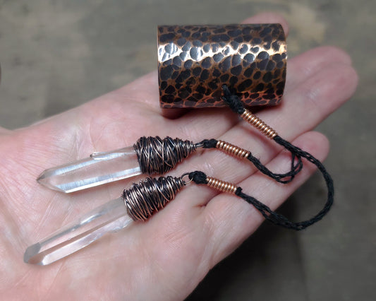 A hand holding a hammered copper hair cuff with two clear crystal points wrapped in copper wire, and black cords against a blurred gray background.