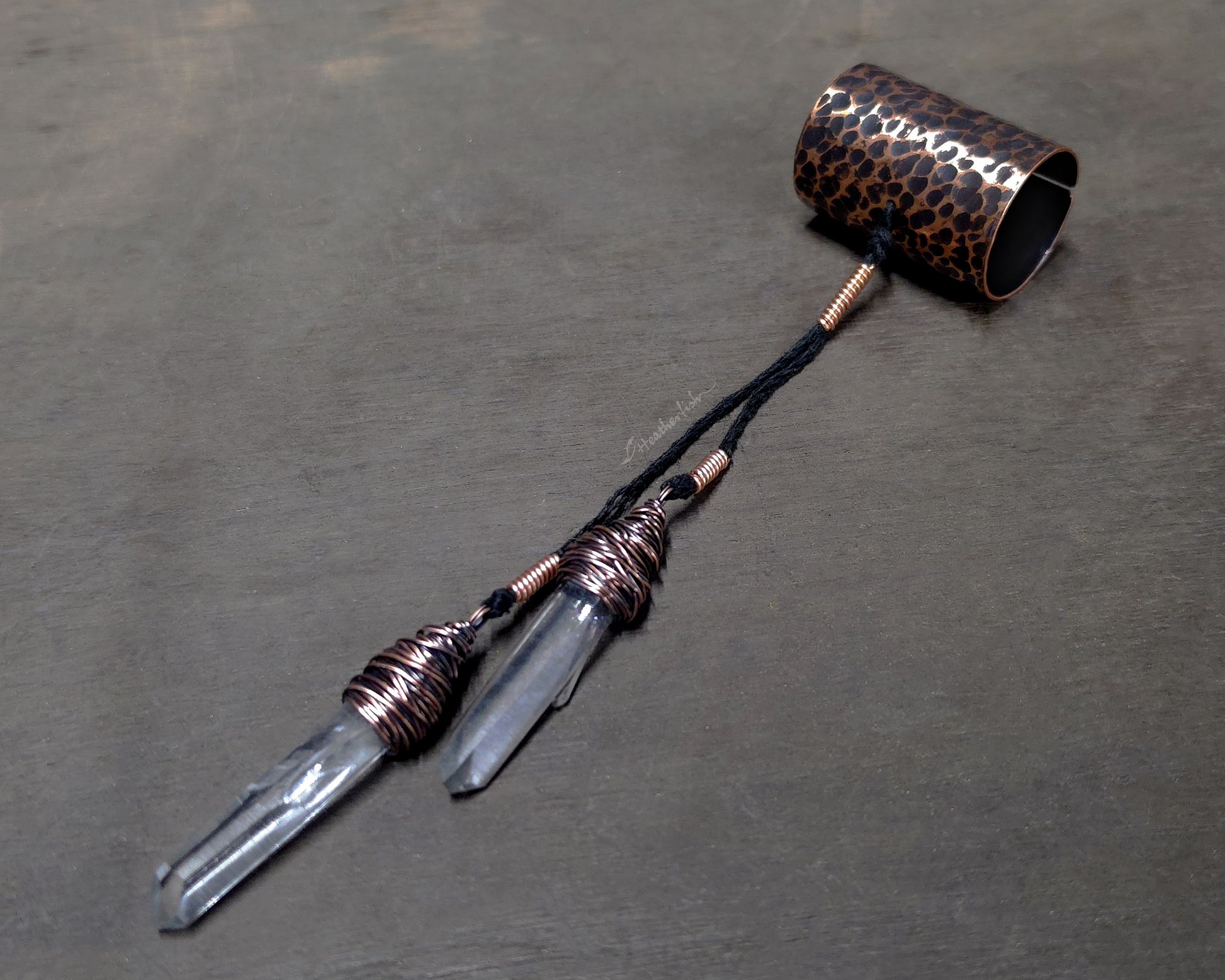 A handmade jewelry piece featuring two clear quartz crystal points wrapped in copper wire, connected by a black cord to a hammered copper cuff displayed on a dark gray textured surface.