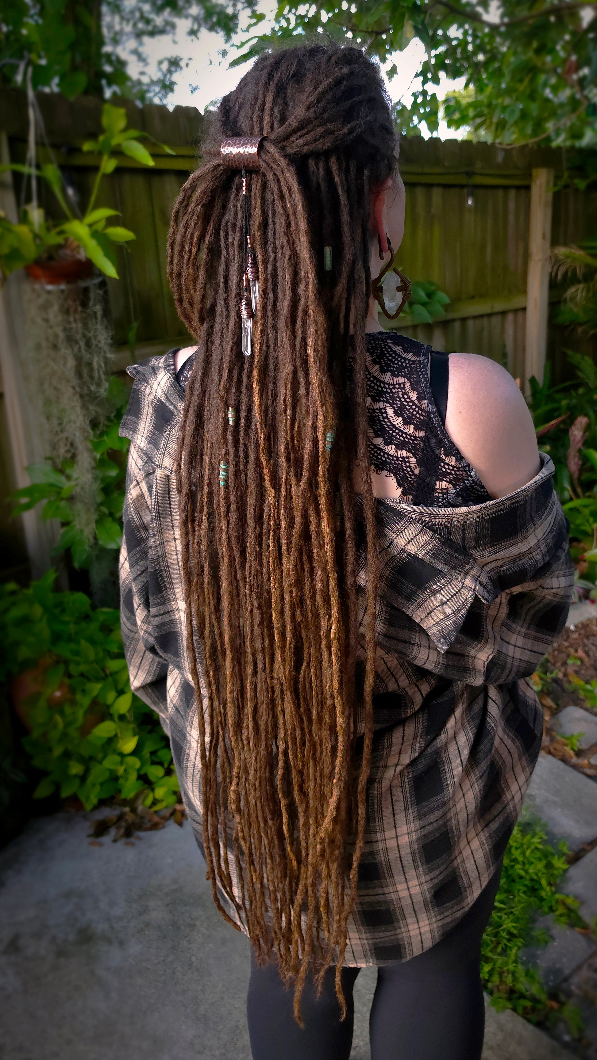 A person shown from the back outdoors, wearing a long brown dreadlock hairstyle with a half-up top knot and a small hair accessory. The person is wearing a copper ear weight, and an off-the-shoulder plaid flannel shirt over a black lace top, set against a backyard fence and garden background.