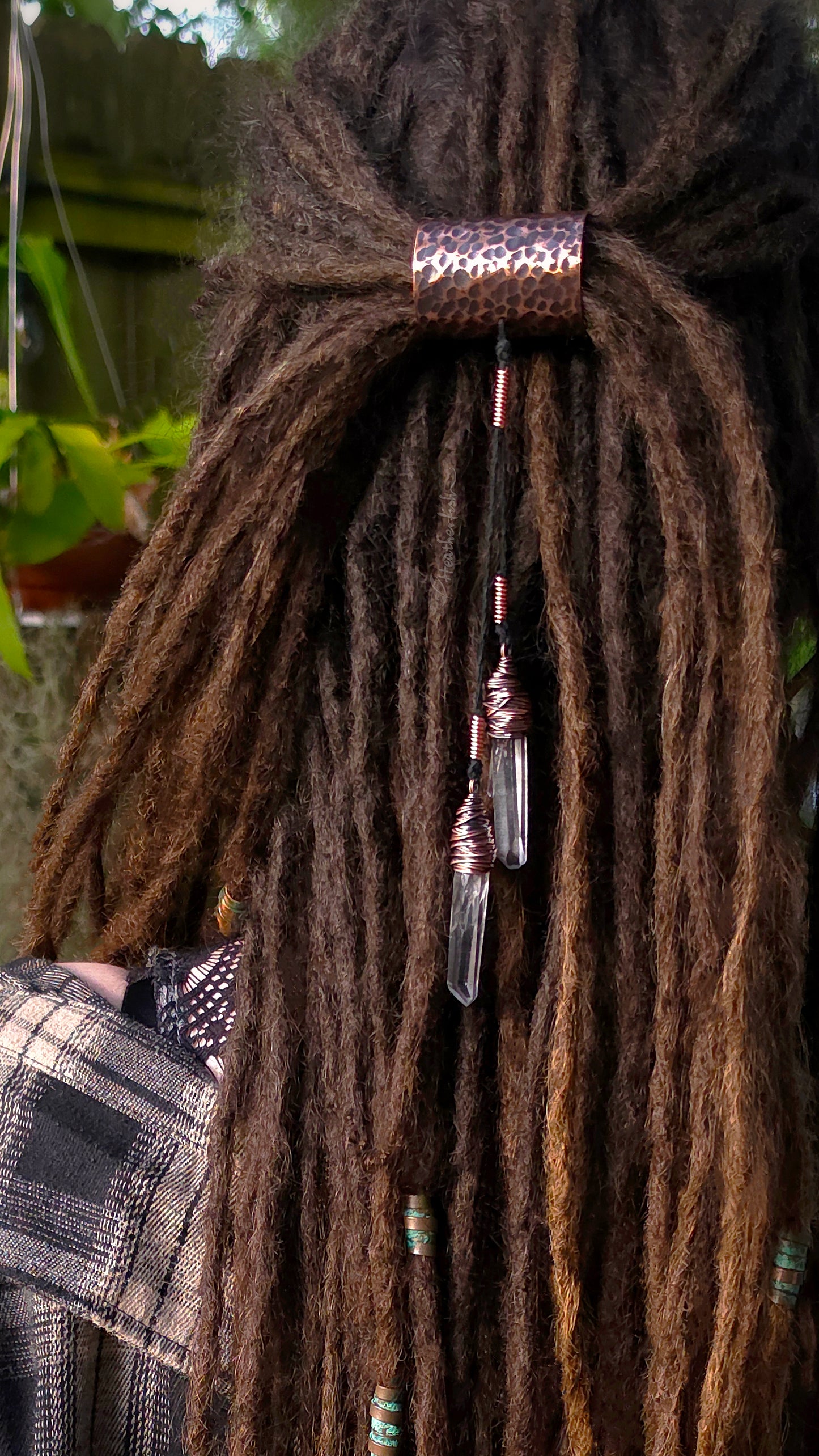 Close-up of a person's dark brown dreadlocks secured with a hammered copper cuff with dangling crystal points, with a plaid shirt visible at the side.