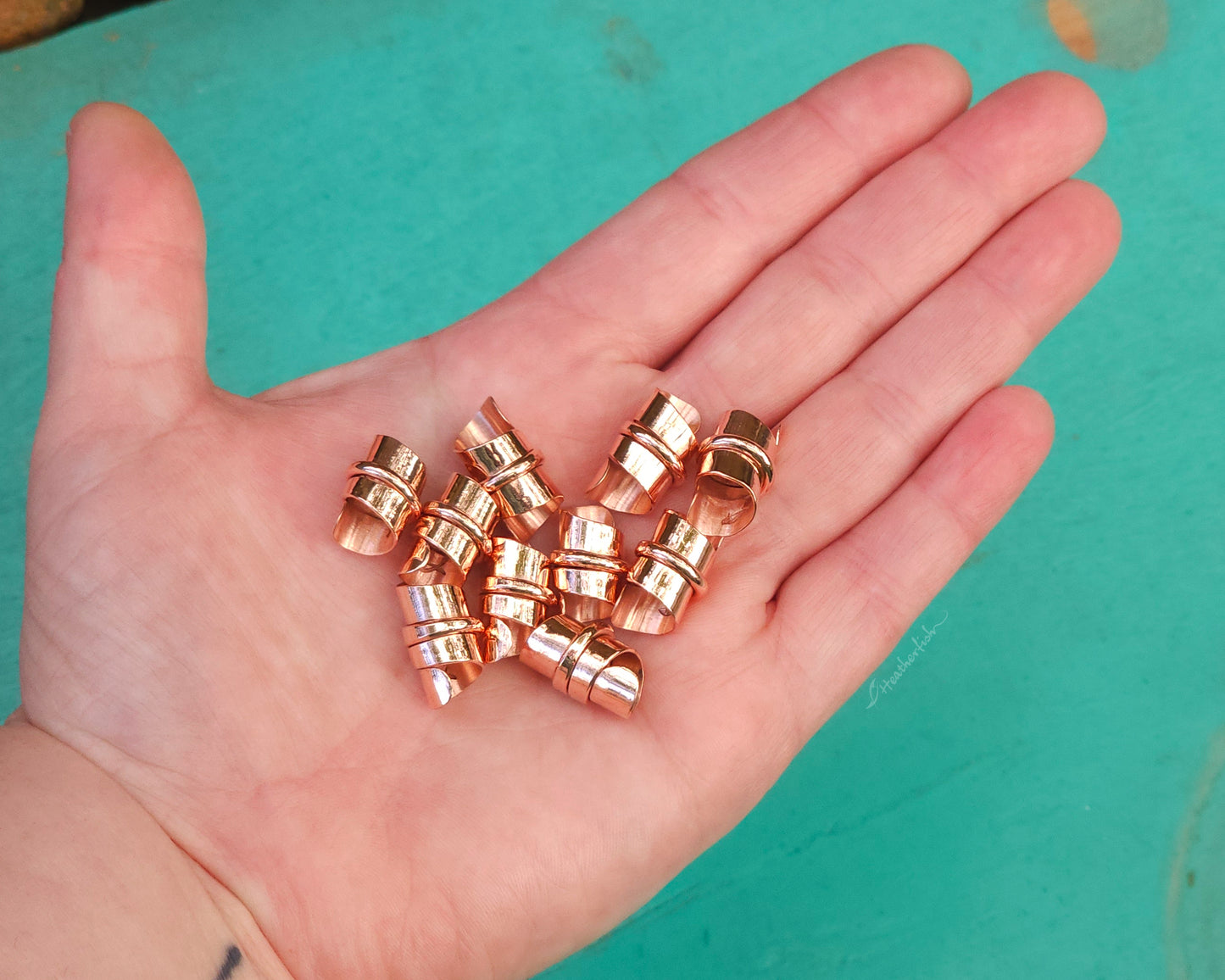 Copper hair jewelry for locs, dreadlocks, braids, or beards, stylized with a center ring and flattened ends, held in the artist&#39;s hand to show scale.