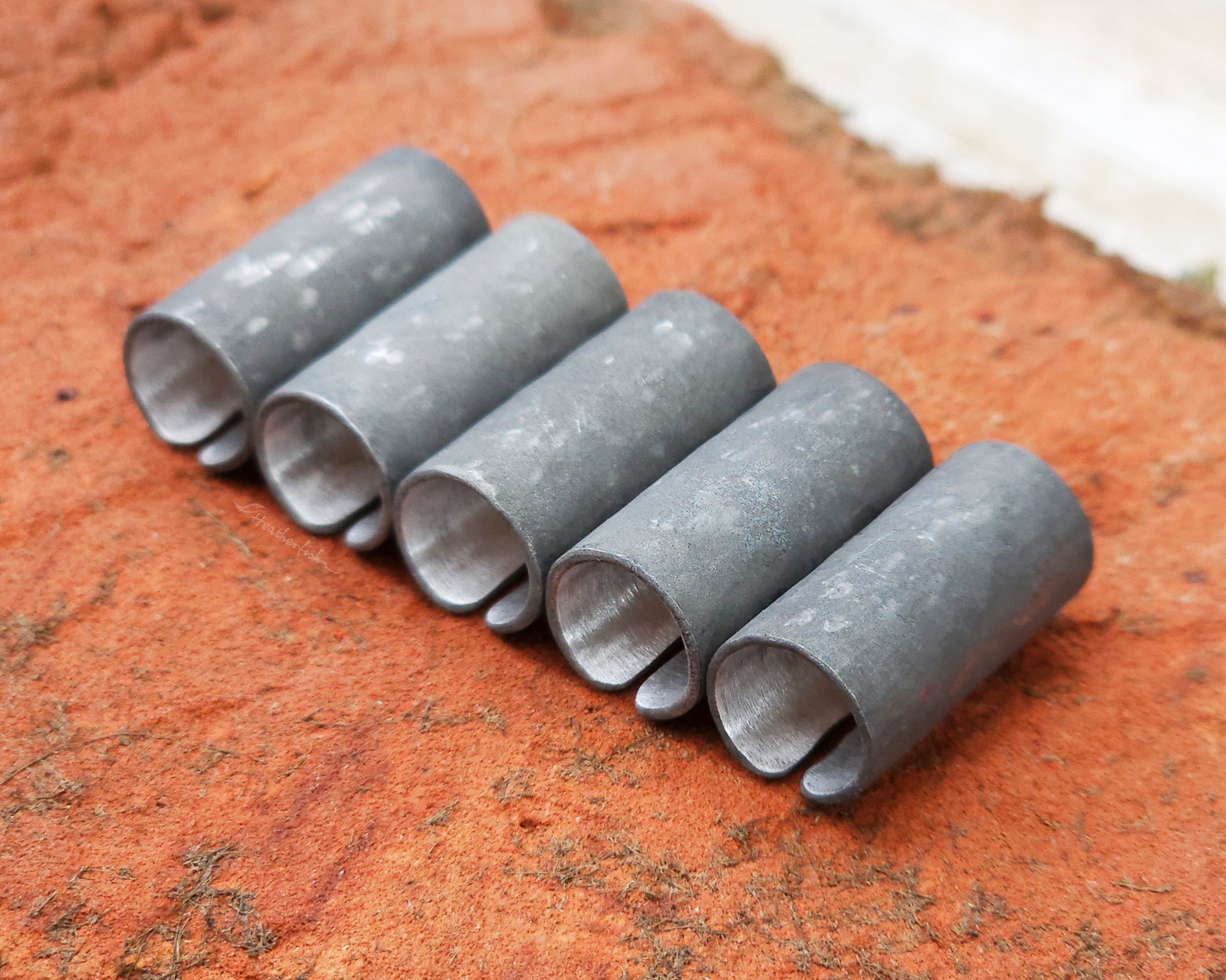 Set of 5 textured oxidized aluminum hair cuffs for locs, dreadlocks, braids, or beards, in a matte grey. Sitting on a reddish orange brick lined up in a row.