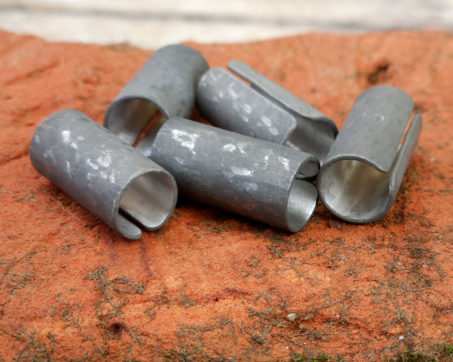 Set of 5 textured oxidized aluminum hair cuffs for locs, dreadlocks, braids, or beards, in a matte grey. Sitting on a reddish orange brick lined up in a random pile.