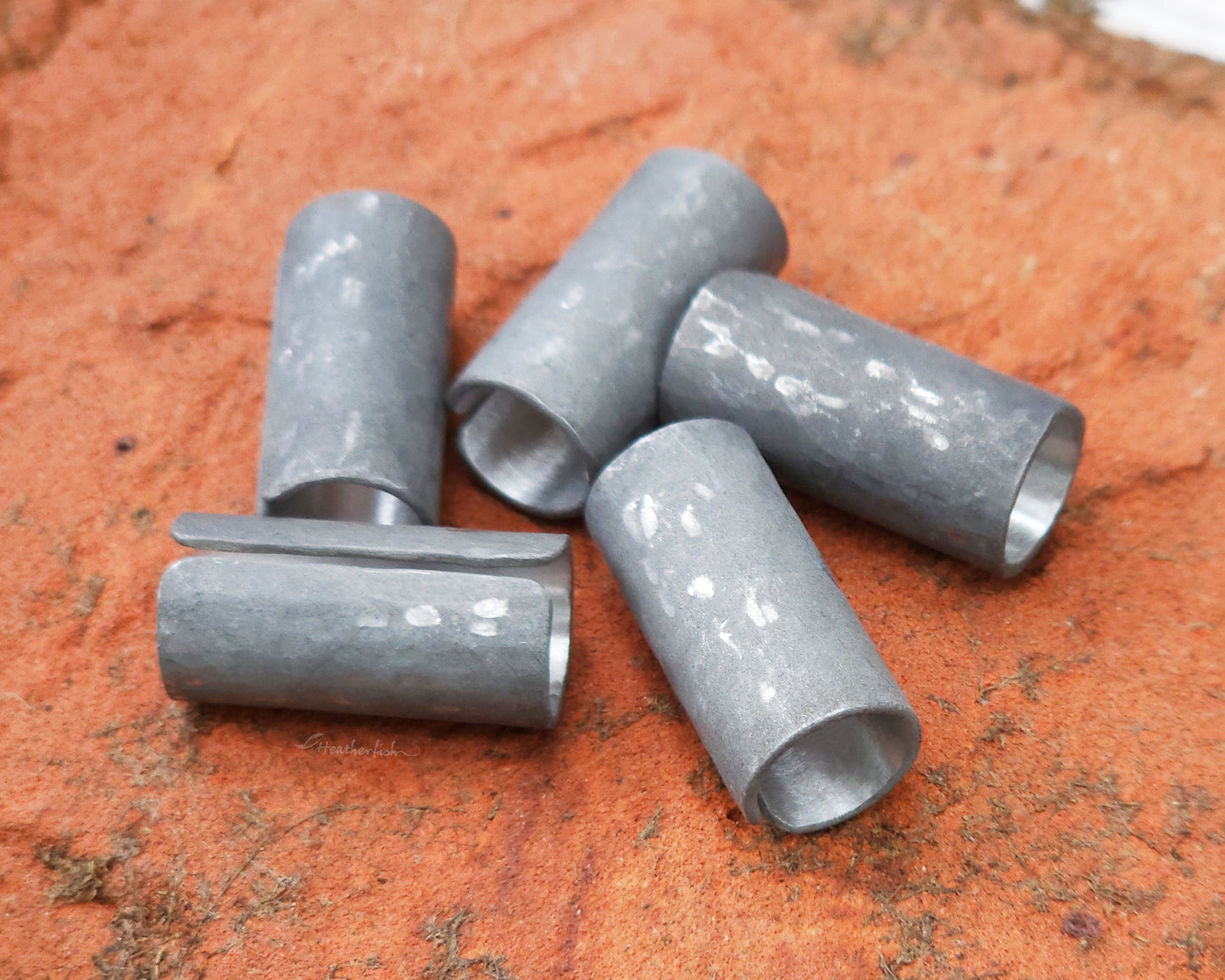 Set of 5 textured oxidized aluminum hair cuffs for locs, dreadlocks, braids, or beards, in a matte grey. Sitting on a reddish orange brick lined up in a random pile.