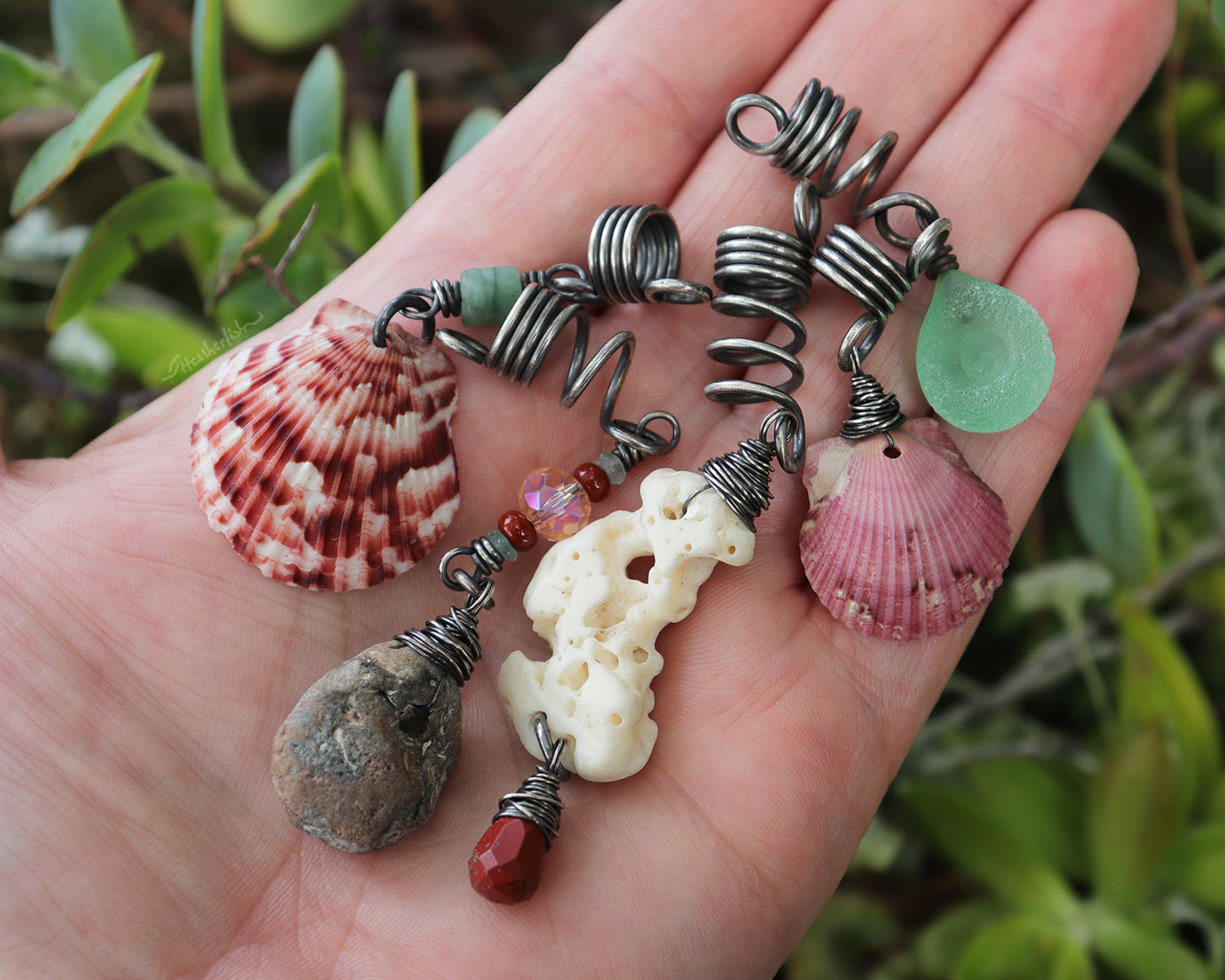 A set of 5 hair beads on silver coils featuring pink shells, coral, red jasper, sea green lampwork glass, a sea stone, and aquamarine, and held in hand to show scale.