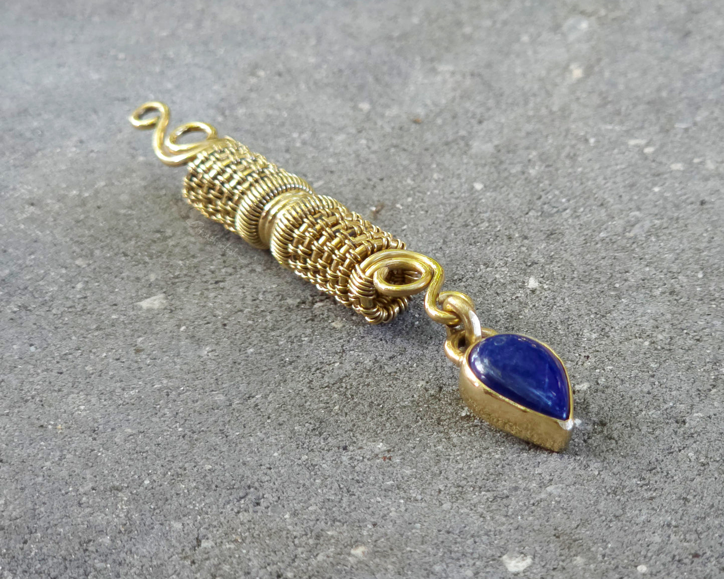 A handmade gold tone wire-wrapped hair bead with a teardrop-shaped blue lapis lazuli stone, displayed diagonally on a textured gray concrete surface.