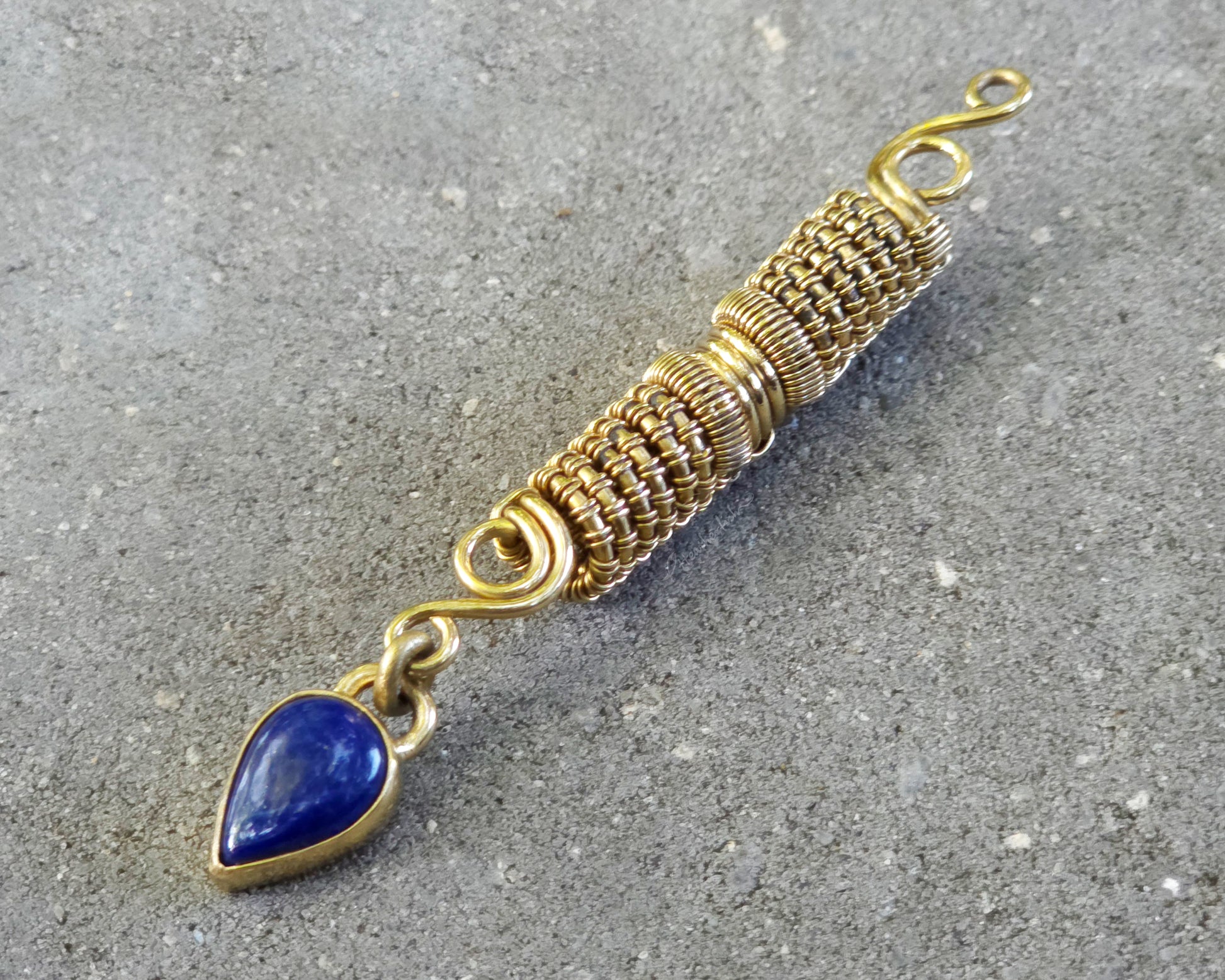 A handmade gold tone wire-wrapped hair bead with a teardrop-shaped blue lapis lazuli stone, displayed diagonally on a textured gray concrete surface.