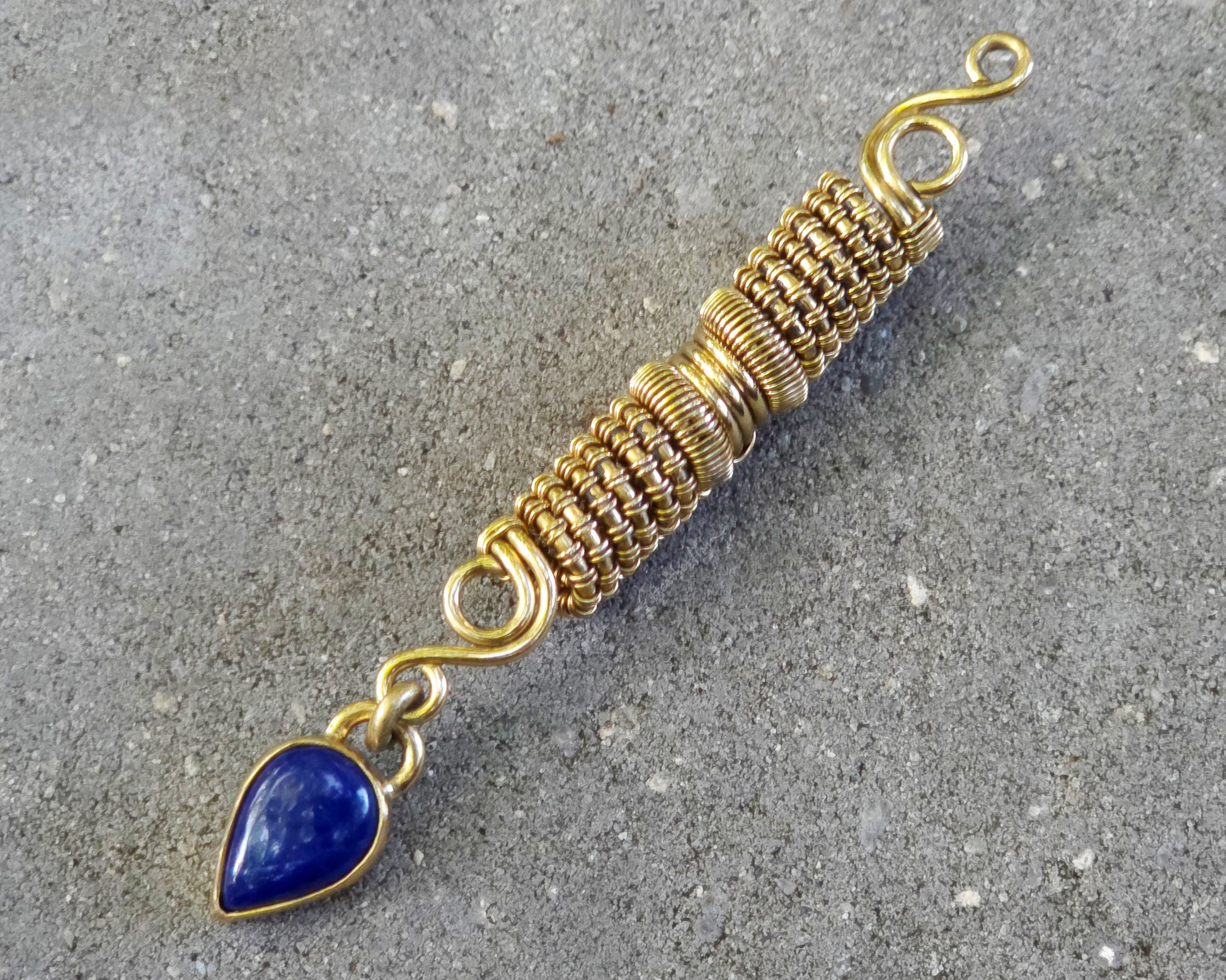 A handmade gold tone wire-wrapped hair bead with a teardrop-shaped blue lapis lazuli stone, displayed diagonally on a textured gray concrete surface.