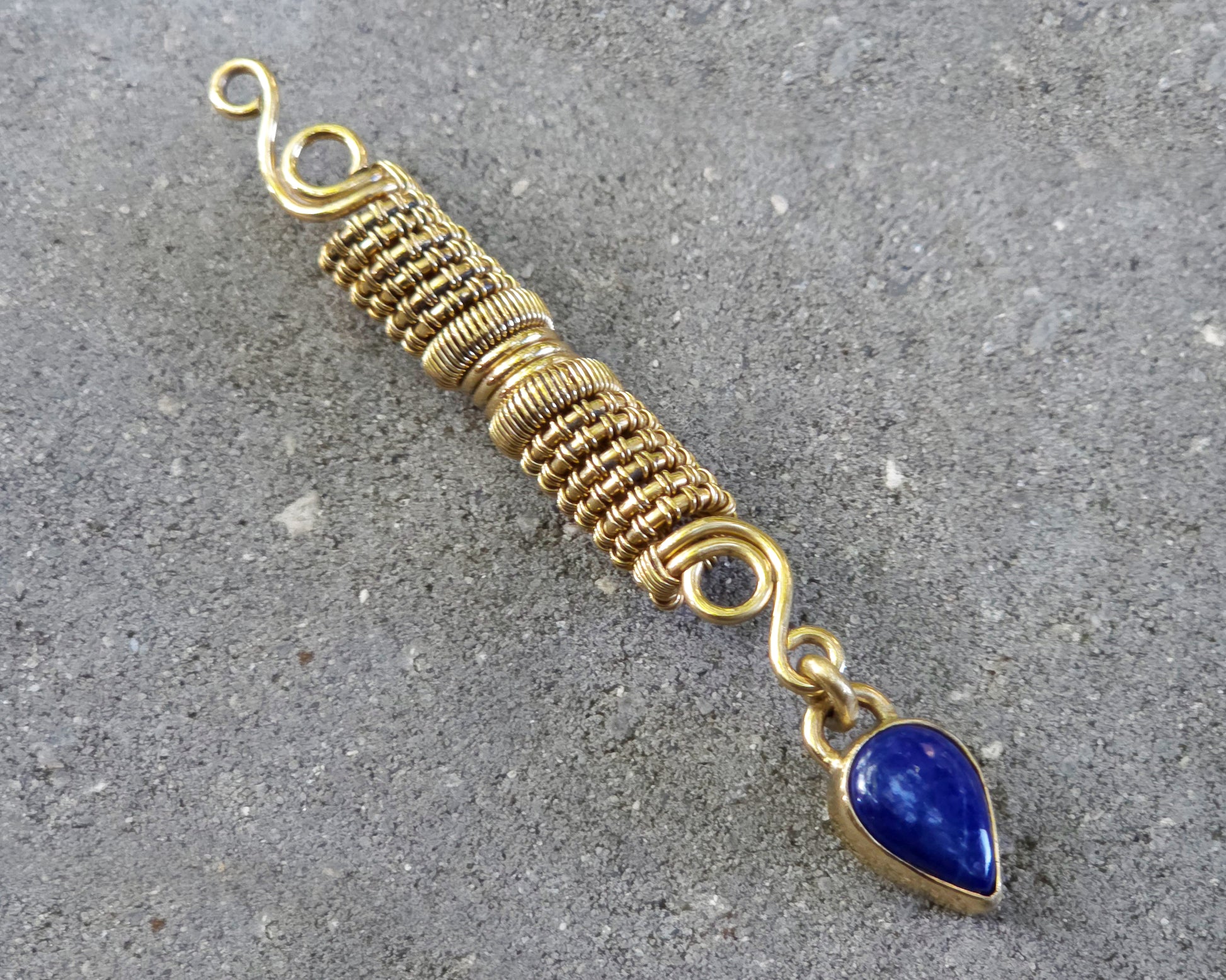 A handmade gold tone wire-wrapped hair bead with a teardrop-shaped blue lapis lazuli stone, displayed diagonally on a textured gray concrete surface.