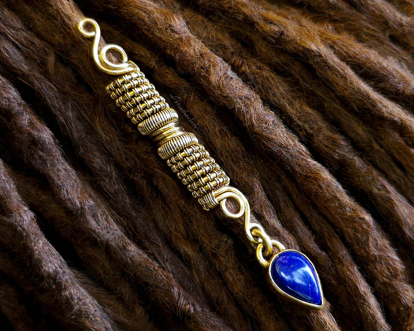 A handmade gold tone wire-wrapped hair bead with a teardrop-shaped blue lapis lazuli stone, displayed diagonally on a brown dreadlock.