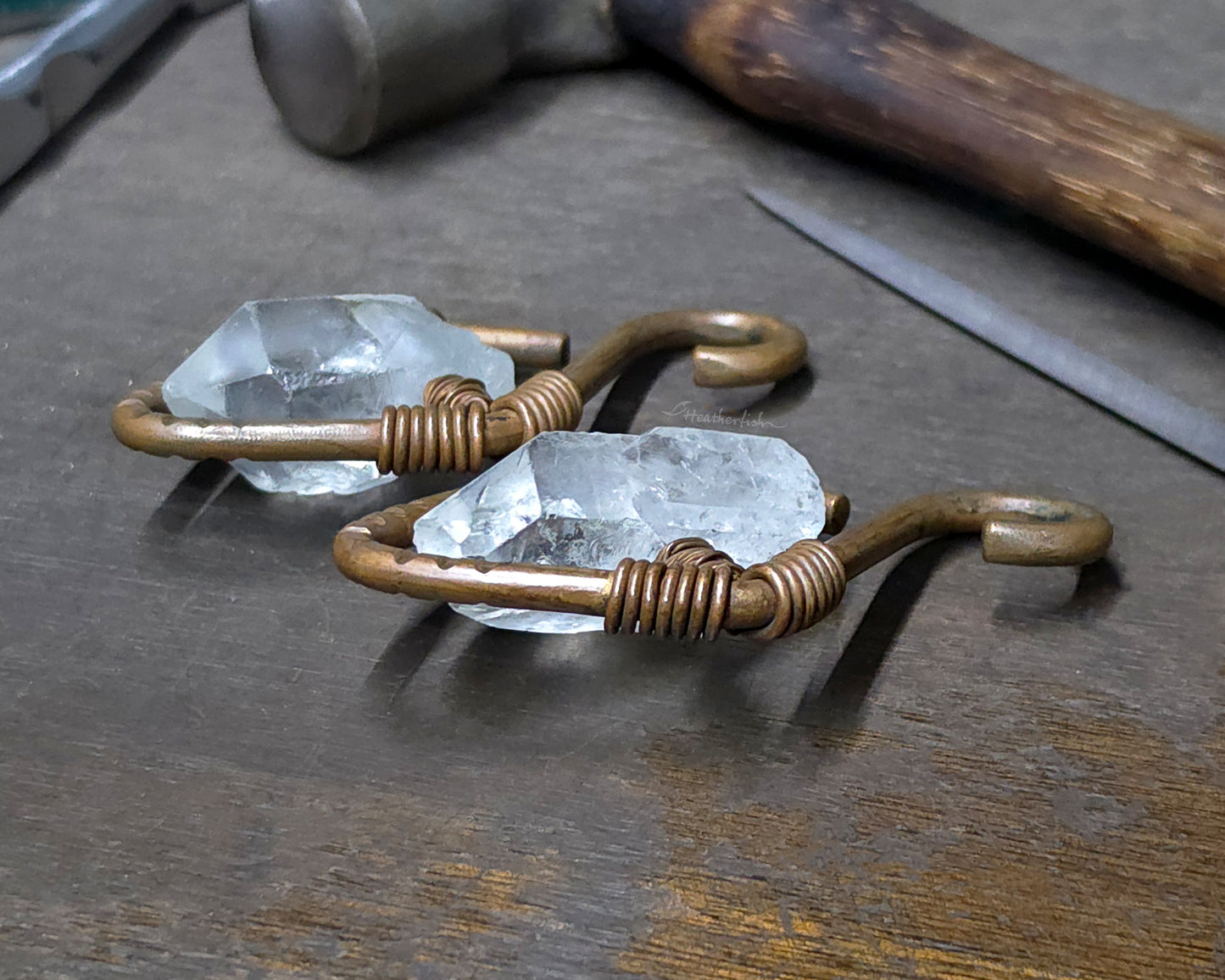 A pair of handmade copper ear weights with clear quartz points displayed on a dark wooden workbench with tools like a hammer and file blurred in the background.