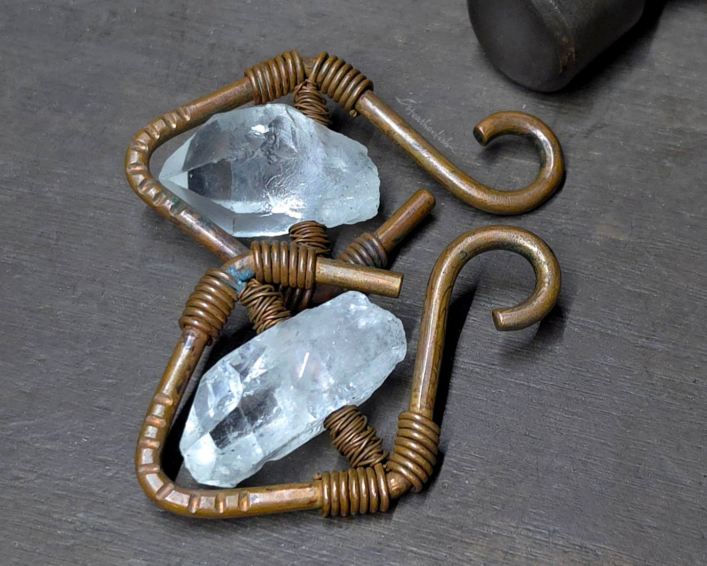 Two handmade copper ear weights with clear quartz crystals and curved copper hooks, displayed on a dark textured surface with a hammer in the background.
