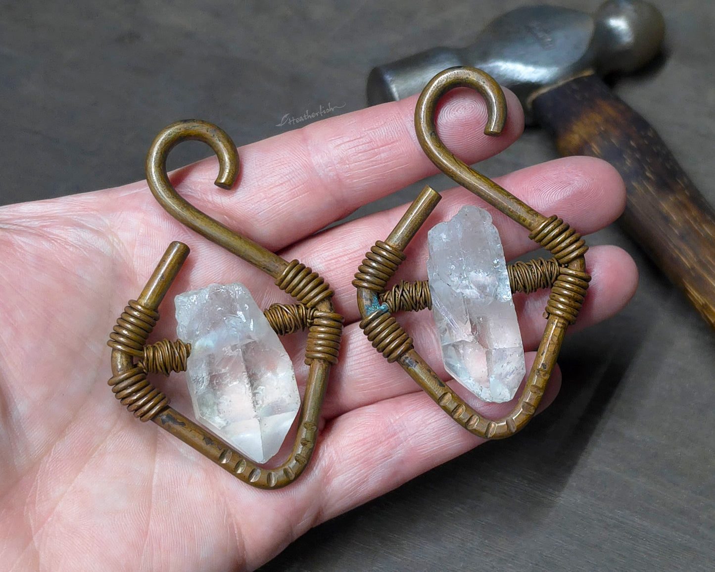 A pair of handmade copper ear weights with clear quartz crystal points held in a person's hand with a hammer blurred in the background.