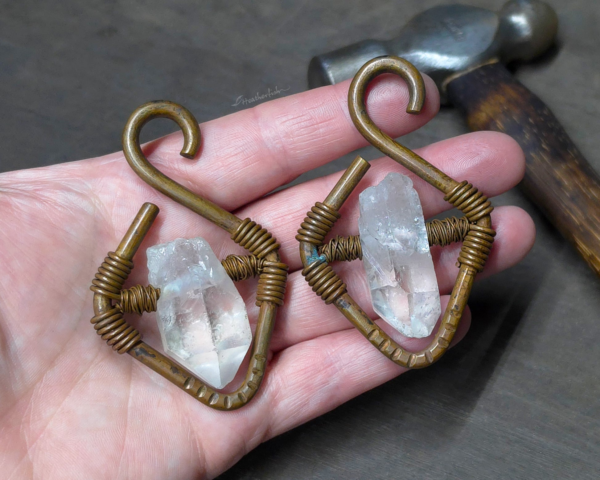 A pair of handmade copper ear weights with clear quartz crystal points held in a person's hand with a hammer blurred in the background.
