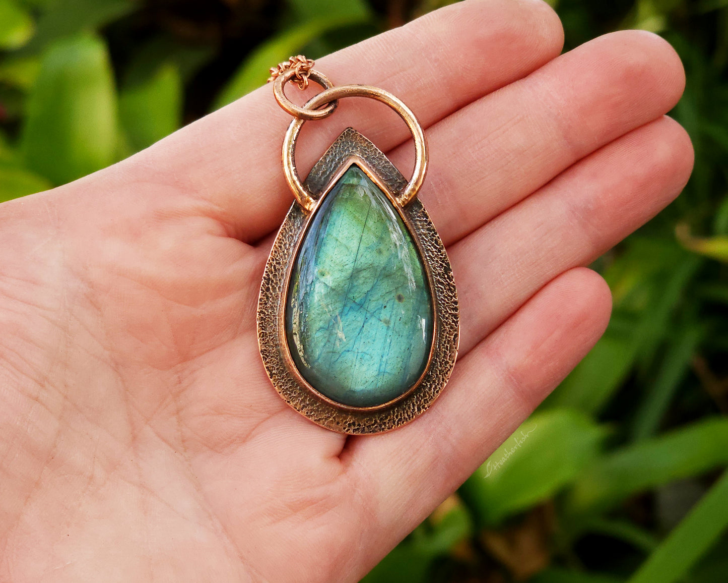 Teardrop-shaped pendant with a green labradorite gemstone held in a hand against a blurred green background