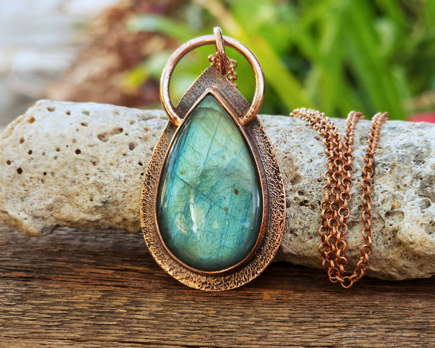 An aqua green pear shaped labradorite pendant set in a copper bezel, propped upright against a rock on a wood board.