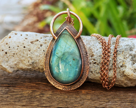 An aqua green pear shaped labradorite pendant set in a copper bezel, propped upright against a rock on a wood board.