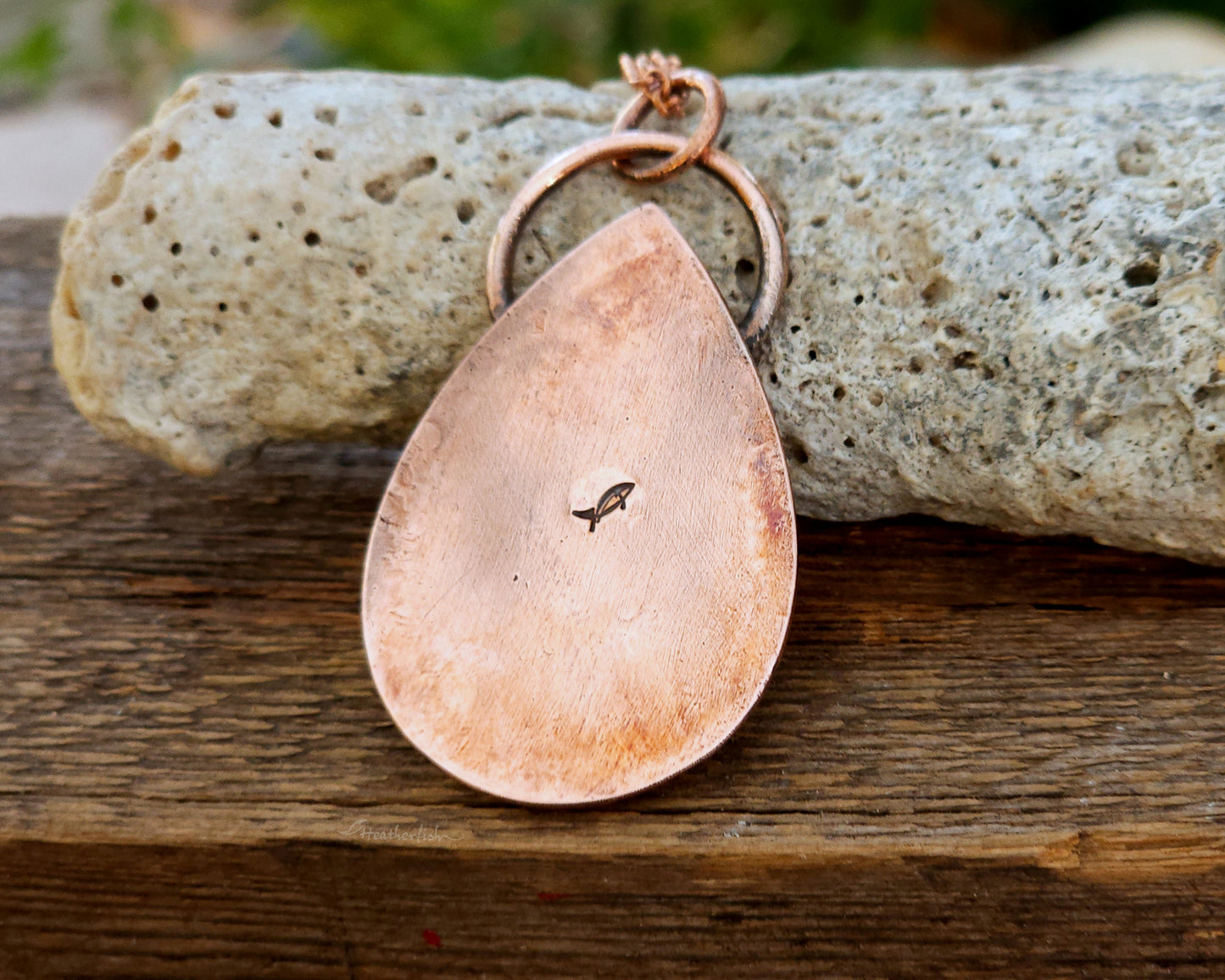 The back of a teardrop-shaped copper pendant on a wooden surface with a blurred natural background