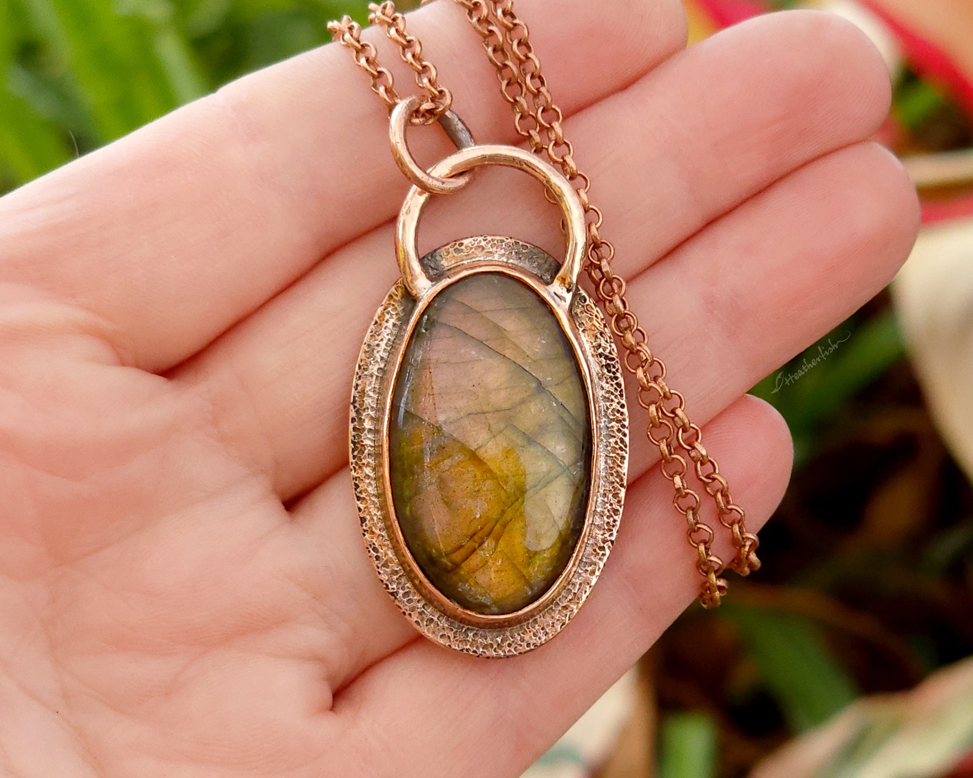 Copper and labradorite pendant held in a hand against a blurred natural background