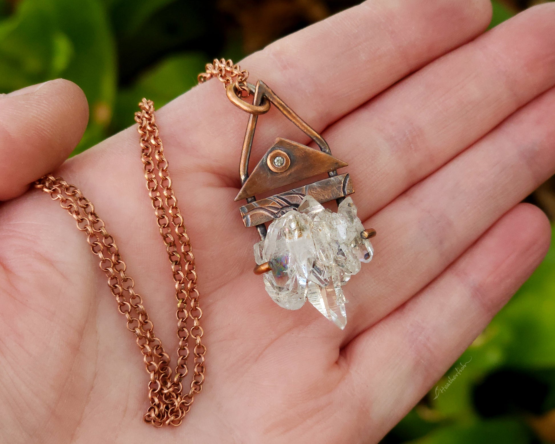 Hand holding a copper chain necklace with a crystal pendant against a blurred green background
