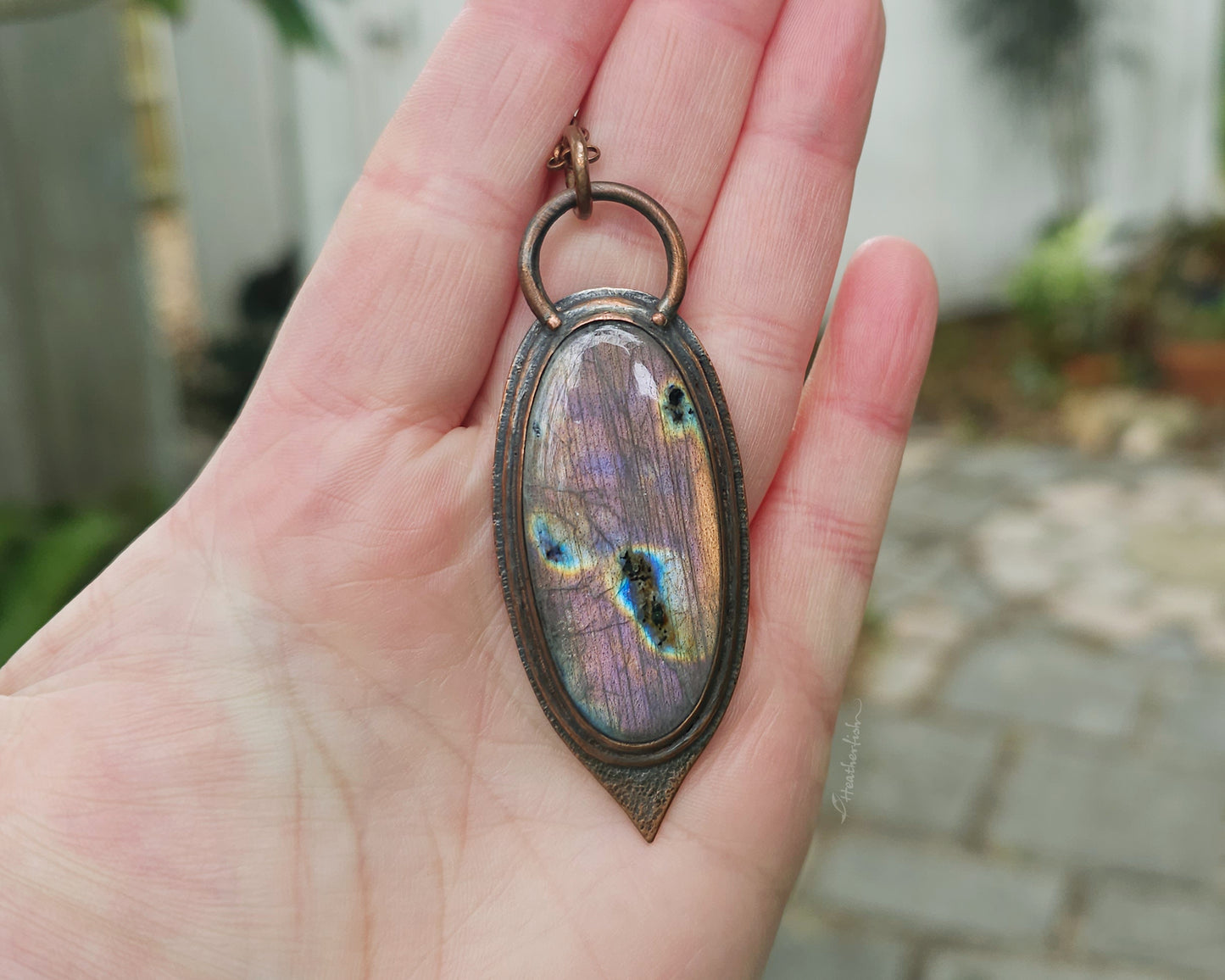 Hand holding a pendant with a colorful stone, blurred outdoor background