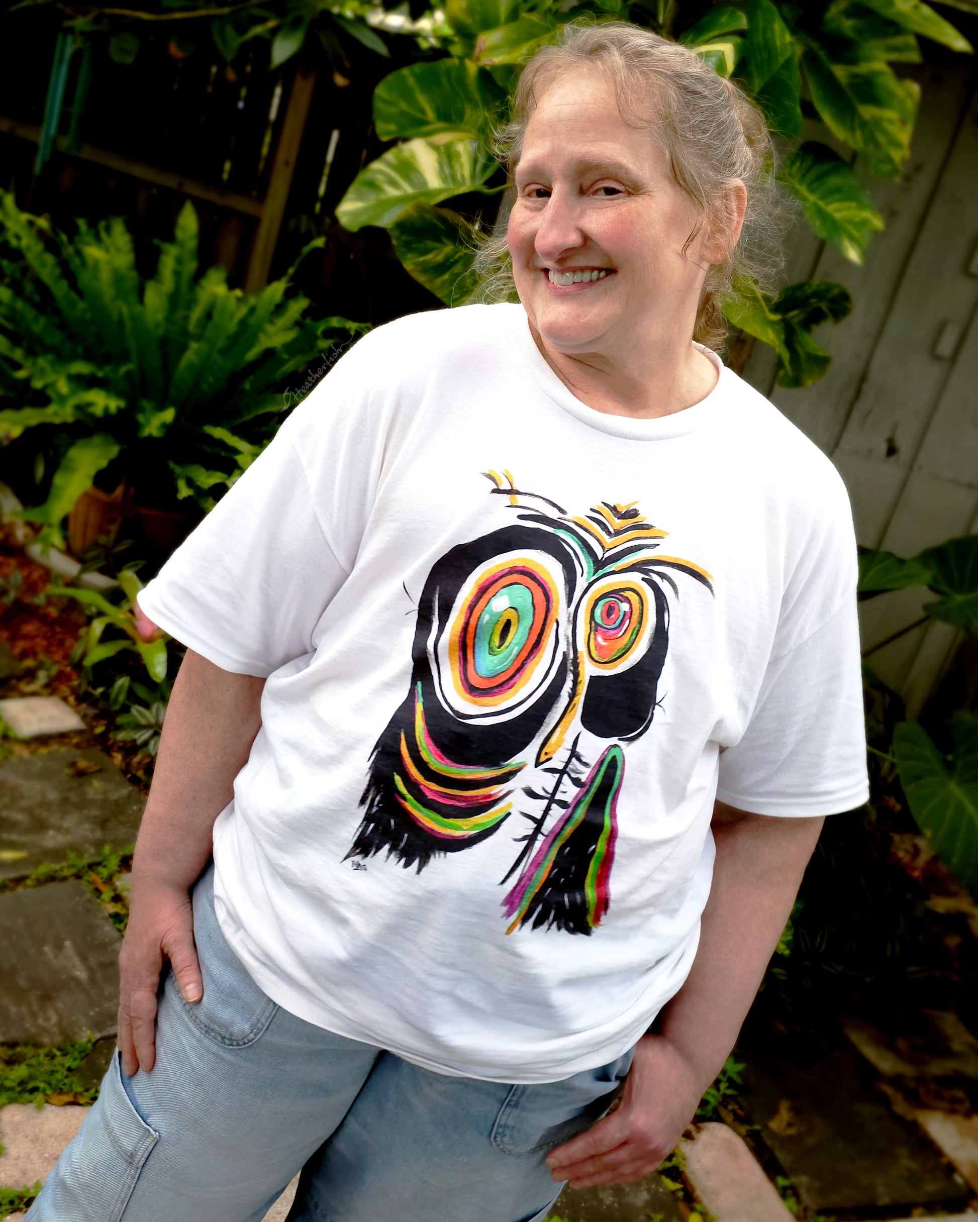 A smiling woman wearing the white sad penguin artisan t-shirt and a pair of jeans while standing on a garden pathway.