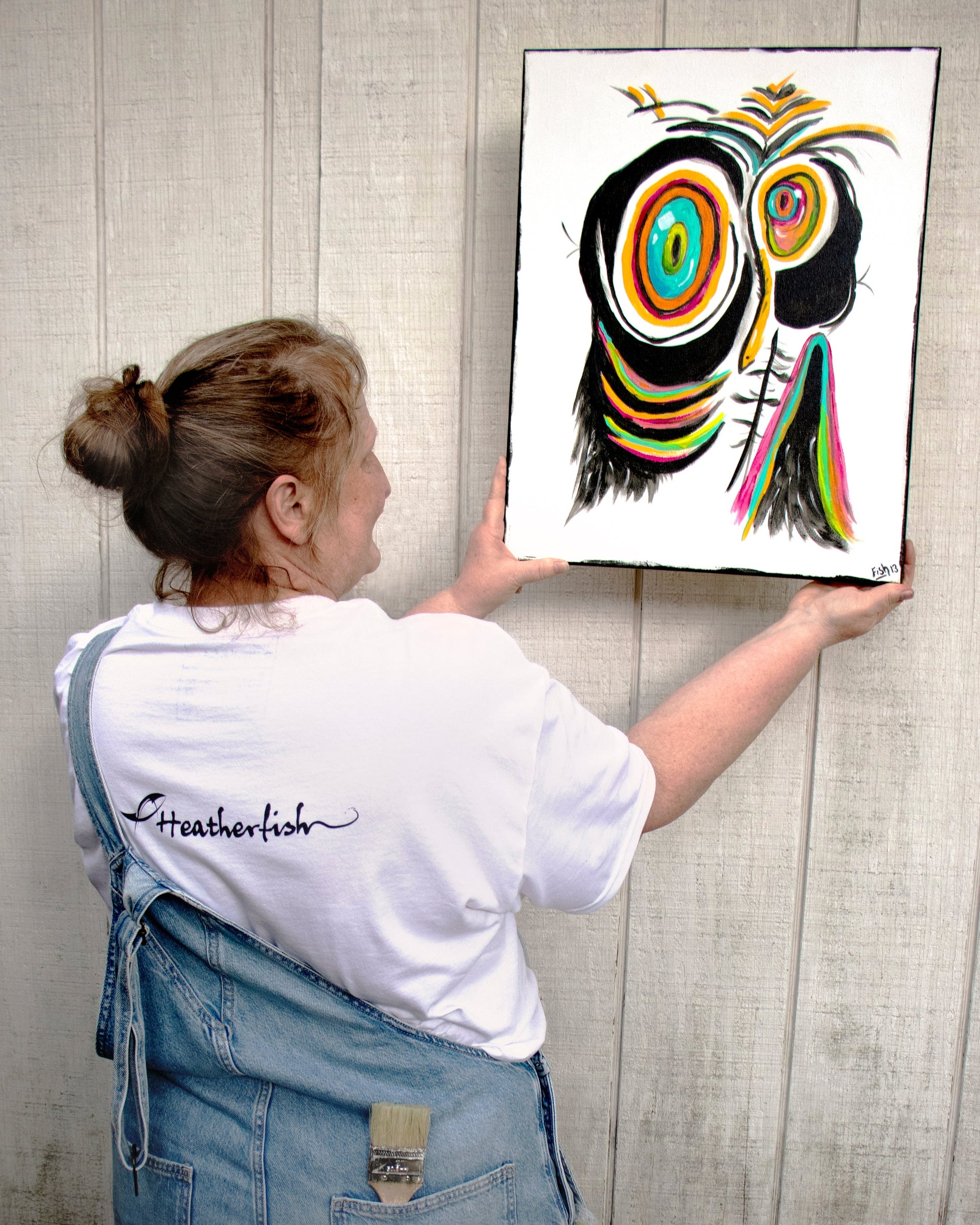 A woman with her back to the camera in a pair of overalls showing the heatherfish logo on the back of the sad penguin white t-shirt holding the original painting of the sad penguin against a rustic wood wall.
