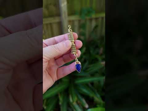 A handmade gold tone wire-wrapped hair bead with a teardrop-shaped blue lapis lazuli stone, held by a hand against a natural background.